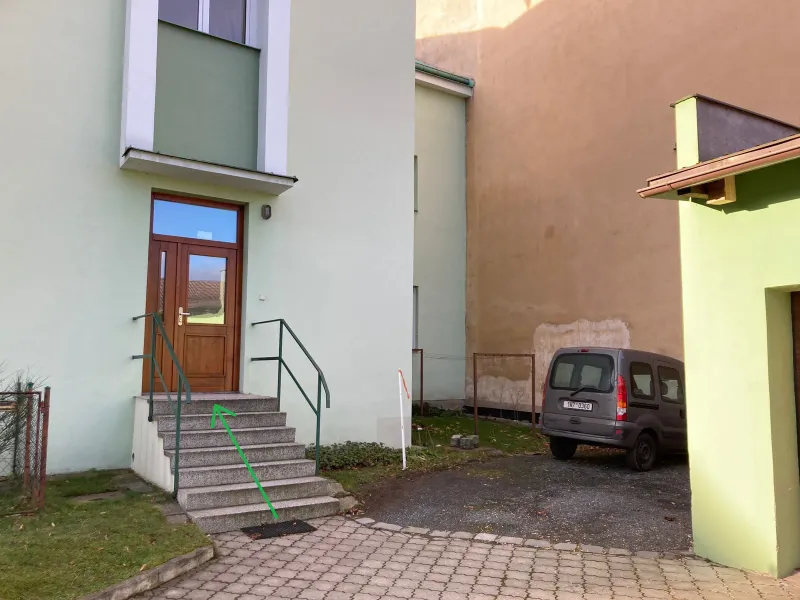 Dřevěné dveře na konci několika betonových schodů se zábradlím, na dlážděném dvoře. Zelená šipka vede po schodech ke dveřím.