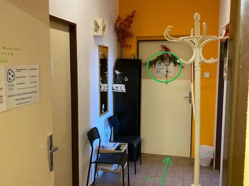 Čekárna s věšákem, židlemi a stolkem s letáky; zelená šipka po podlaze vede ke dveřím v pozadí.