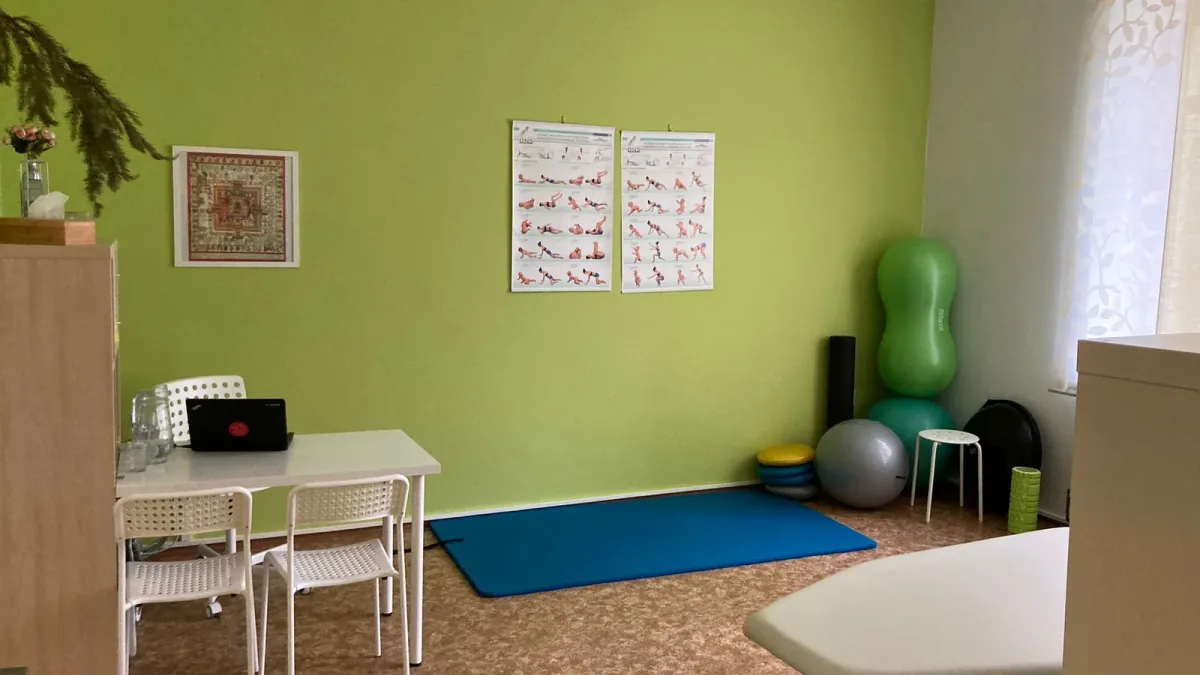 Terapeutická místnost se zelenou stěnou: stůl se židlemi, nákresy cviků na stěně, v rohu gymnastické míče a podložky, vpravo lehátko.