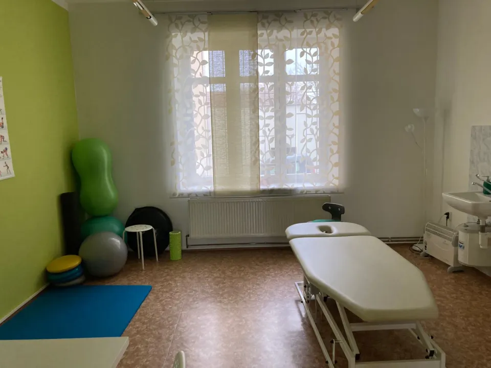 Terapeutická místnost s velkým oknem: polohovací lehátko, umyvadlo, v rohu gymnastické míče, válec a podložky.