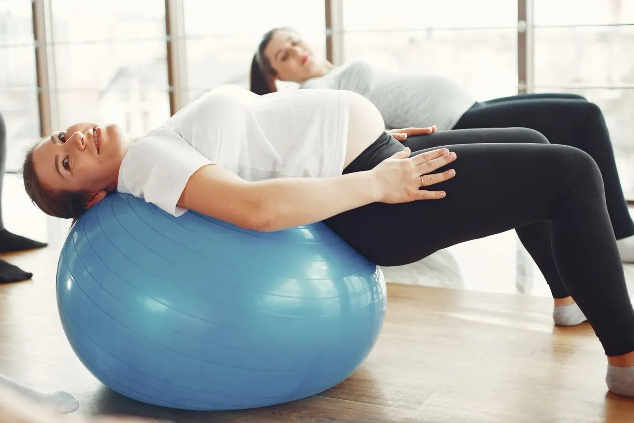 Těhotná žena leží v záklonu přes gymnastický míč, v pozadí cvičí druhá žena