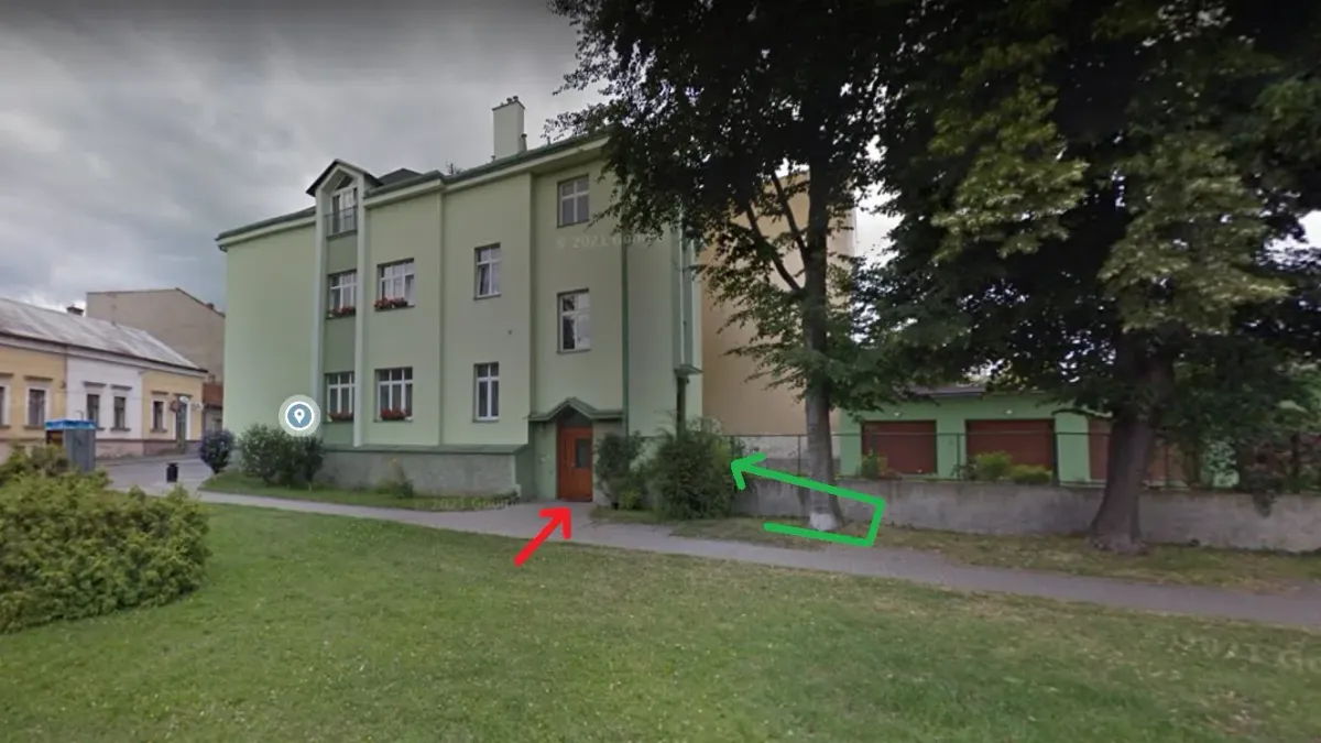 Světle zelený patrový dům, ve kterém centrum sídlí, při pohledu přes trávník. Červená šipka ukazuje na dveře v čelní stěně, zelená šipka vede kolem rohu domu dozadu.